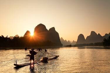 China, Guangxi Zhuangzu Zizhiqu, Guangxi, Yangshuo, Cormorant fisherman on the Li River at sunset, Karst mountains in the background (Luigi Vaccarella) - Muzeo.com
