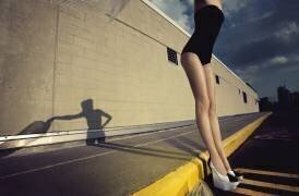 Femme en maillot de bain une pièce et chaussures à talons hauts debout contre une bordure de trottoir (anonyme) - Muzeo.com